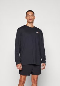 Must musta-sleeve särk ümmarguse kaelusega ja UA logo oranžina rinnal, koos mustade lühikeste pükstega. Sujuv tekstuur, vabaaja lõige.