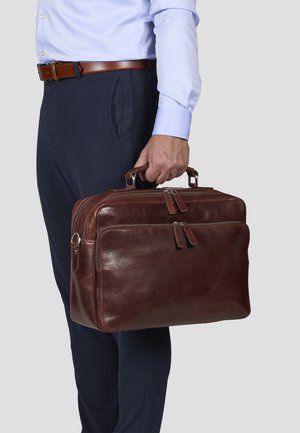 Brown leather briefcase with double zippers, top handles, and a smooth texture. Held by a person wearing navy pants and a light blue shirt.