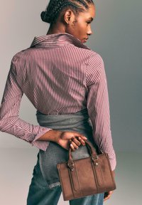 Brun læderhåndtaske med små top-håndtag, rektangulær form, synlige sømme, holdt i en hånd. Modellen bærer en rød og hvid stribet skjorte.