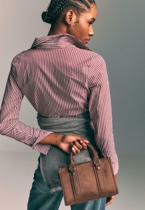 Borsa a mano in pelle marrone con piccole maniglie superiori, forma rettangolare, cuciture visibili, tenuta in un mano. La modella indossa una camicia a righe rossa e bianche.