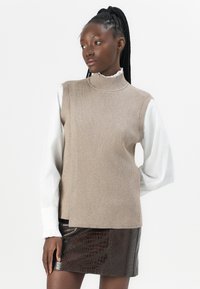 Haut sans manches beige à côtes avec un col haut, superposé sur une chemise blanche à manches longues à volants, associé à une jupe brillante en cuir de crocodile marron.