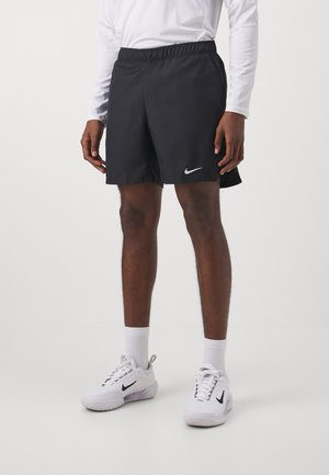 Mężczyzna w czarnych sportowych szortach Nike, białej koszulce z długim rękawem, białych butach sportowych Nike i białych skarpetach, stojący na białej podłodze.
