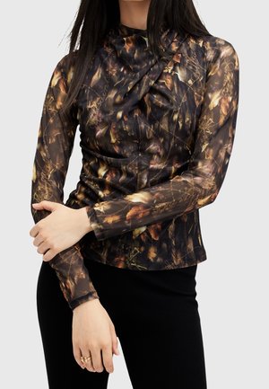 Kvinde iført en langærmet, tætsiddende top med mørkt blomstermønster og kædeprint, kombineret med sorte bukser, stående foran en ensfarvet baggrund.