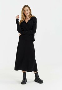 Zwarte gebreide trui met kraag, gecombineerd met een lange zwarte rok. Model draagt stevige zwarte laarzen. Gladde textuur, minimalistisch design.