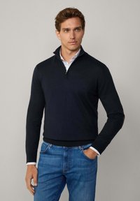 Marinblå zip-up tröja med långa ärmar, ribbade manschetter och hög krage. Inkluderar kontrasterande bruna armbågsfickor. Bärs med ljusblå jeans.