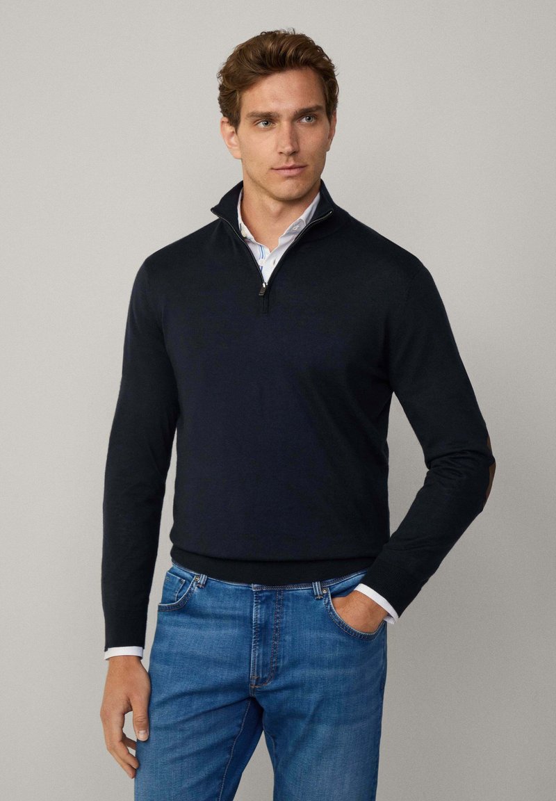 Marinblå zip-up tröja med långa ärmar, ribbade manschetter och hög krage. Inkluderar kontrasterande bruna armbågsfickor. Bärs med ljusblå jeans.