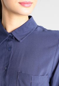 Mörkblå skjorta med krage i ett slätt tyg, med knapprad och bröstficka. Kragen är strukturerad med subtila sömmar.