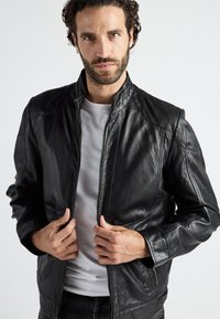 Zwarte leren jas met ritssluiting, gestructureerde schouderpanelen en een staande kraag. Glad afwerking, getailleerd ontwerp en lange mouwen.