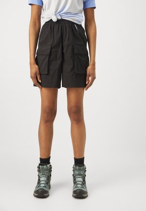 Persona que lleva pantalones cortos cargo negros, camisa anudada azul claro, calcetines negros y botas de senderismo grises sobre un fondo blanco.