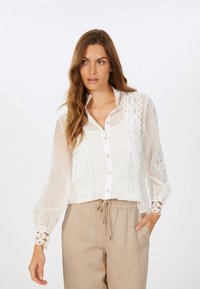 Weiße durchsichtige Bluse mit dekorativen Spitzenpaneelen und Knopfleiste. Verfügt über lange Ärmel und Spitzenmanschetten. Kombiniert mit beigefarbenen Hose mit Kordelzug.