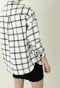 Koszula w białą i czarną kratę z teksturowanej tkaniny, z długimi rękawami, zapinanymi na guziki mankietami oraz lekko zaokrąglonym dołem.
