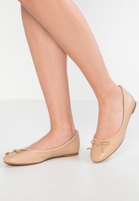 Ballerine in pelle beige con punta arrotondata e dettaglio fiocco, caratterizzate da un tacco basso e una texture liscia su sfondo bianco.