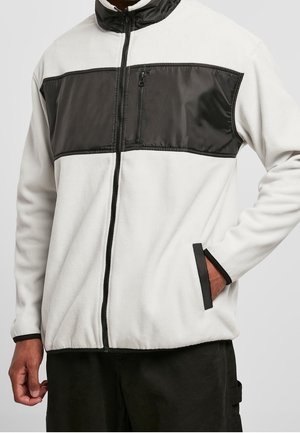 Homme portant une veste en polaire blanche avec une poche poitrine zippée noire et des bordures noires aux poignets et à l'ourlet.