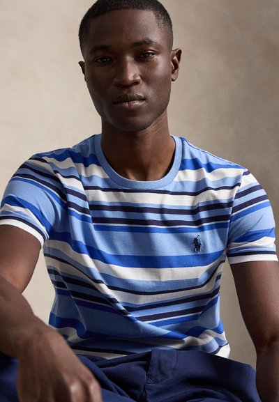 Jeune homme portant une chemise à manches courtes rayée bleue, blanche et bleu marine, assis avec une expression neutre devant un fond uni.