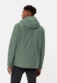 Giacca verde resistente all'acqua con cappuccio, caratterizzata da una texture liscia e un design pulito. Include cuciture laterali e un logo discreto.