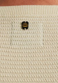 Crème-kleurige gebreide trui met een textuurpatroon en een klein zwart "PME Legend" label met gouden opdruk op de achterkant.