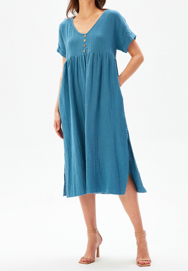 Une robe bleu clair à manches courtes en tissu texturé avec un décolleté en V et trois boutons en bois à l'avant. Elle comporte des fentes sur les côtés.