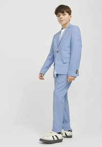 Veste et pantalon de costume bleu clair, avec une fermeture à un bouton, associés à un t-shirt blanc et des chaussures de sport.