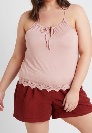 Kobieta nosząca jasnoróżowy bezrękawnik z koronkowym wykończeniem i wiązanym detalem, w połączeniu z luźnymi ciemnoczerwonymi szortami.