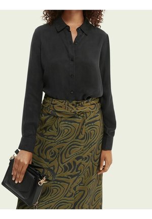 Vrouw die een zwart geknoopt overhemd en een groene rok met hoge taille en zwarte abstracte patronen draagt, met een zwarte handtas in haar linkerhand.