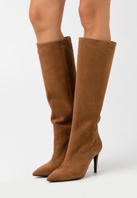 Bruine suède laarzen tot aan de knie met een puntige neus en een naaldhak. Zachte textuur en een nauwsluitend ontwerp, geschikt voor formeel of casual gebruik.
