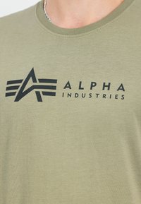Olīvu zaļš kokvilnas T-krekls ar īsām piedurknēm un apaļu kakla izgriezumu. Satur melnu uzdrukātu logotipu "ALPHA INDUSTRIES" ar stilizētiem dizaina elementiem.