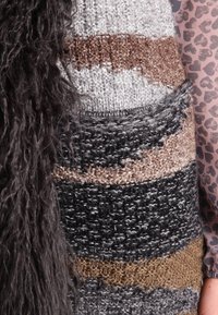 Tessuto di lana lavorato a maglia marrone, grigio e nero con frange lunghe di filato scuro, accanto a un tessuto rosa con cuori scuri, posato sull'avambraccio di una persona.