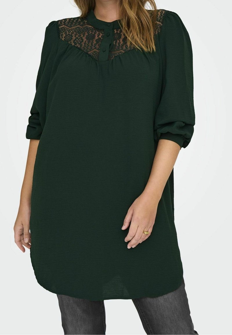 Femme portant une tunique à manches longues vert foncé avec empiècement en dentelle et col boutonné, associée à un jean gris foncé.