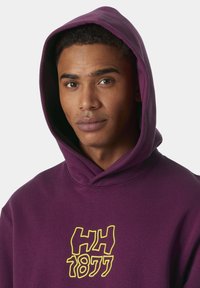 Sudadera púrpura con un gran logo gráfico amarillo que dice "HH 1877" en el pecho, con capucha ajustable y textura suave.