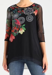 Schwarze Tunika mit floralem Druck, ausgestattet mit roten und grünen Akzenten. Rundhalsausschnitt, dreiviertellange Ärmel und ein fließender Saum. Durchsichtige Textur.