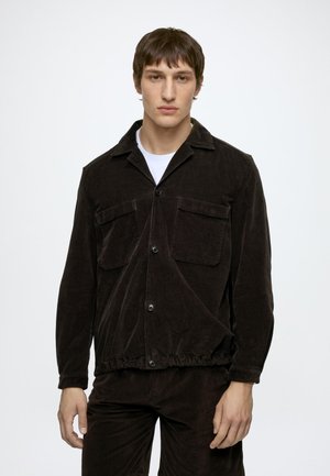 Jeune homme portant une veste marron foncé boutonnée avec deux poches poitrine et un pantalon assorti sur un fond clair uni.