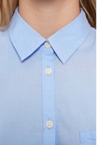 Svetlomodrá bavlnená košeľa s klasickým golierom, šiestimi gombíkmi a malým náprsníkom. Hladká textúra s minimálnymi vráskami.