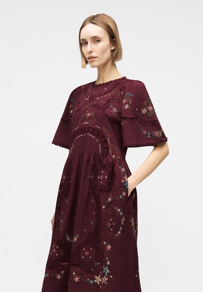 Γυναίκα που φοράει μπορντό φόρεμα με κεντήματα λουλουδιών και λεπτομέρειες από δαντέλα, όρθια με το ένα χέρι στην τσέπη της μπροστά σε απλό φόντο.