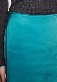 Teal Satinrock mit glatter Oberfläche, der ein elegantes Design und einen leichten Glanz aufweist, kombiniert mit einem schwarzen, strukturierten Langarmoberteil.