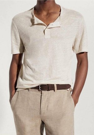 Chemise henley beige à manches courtes avec col et trois boutons, associée à un pantalon crème et une ceinture en cuir marron.