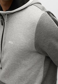Felpa grigia con zip realizzata in tessuto morbido e testurizzato. Presenta un cappuccio nero, un logo distintivo sul petto e pannelli delle maniche a contrasto.