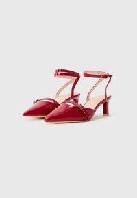 Red patent leather slingback heels with a pointed toe and a mid-height block heel. Features an ankle strap and decorative buckle detail.