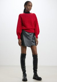 Pullover a maglia rossa con texture e maniche a palloncino, abbinato a una gonna avvolgente in pelle nera e stivali alti fino al ginocchio neri e lucidi. Sfondo semplice.