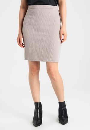 Frau trägt einen beigen knielangen Rock mit Struktur, ein schwarzes Oberteil und schwarze spitze Stiefeletten. Sie steht mit entspannten Händen an den Seiten.