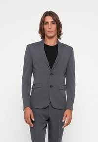 Grey tailored suit jacket with notched lapels, two buttons, and two front pockets, worn over a black t-shirt. Smooth texture, modern fit.