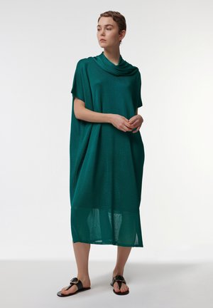 Robe en tricot verte avec une coupe ample, décolleté drapé, longueur midi, et un motif texturé. Associée à des sandales plates noires.