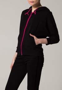 Felpa nera con zip, dotata di cordini rosa, tasche anteriori e una vestibilità morbida. Tessuto soffice e cappuccio per un calore aggiuntivo.