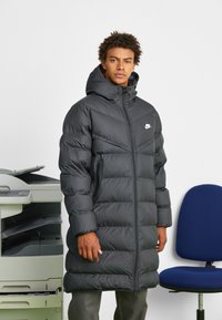 Crni puffer kaput s kapuljačom, s plaštem od šivaćih dijelova, prednjim patentnim zatvaračem i bočnim džepovima; akcent logotipa na prsima.