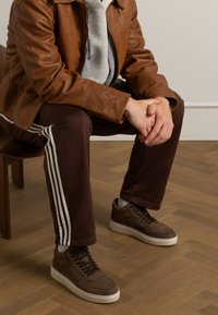 Braune Lederjacke über einem grauen Hoodie, kombiniert mit braunen Hosen mit weißen Streifen. Braune Sneakers mit weißen Sohlen. Hintergrund aus Holzfußboden.