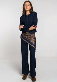 Chaqueta vaquera con mangas abullonadas y botones redondos, combinada con pantalones de pierna ancha y una bufanda a rayas marrón y azul marino usada como cinturón.