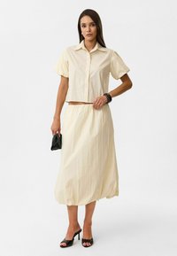Crème-kleurig, cropped button-up shirt met pofmouwen, gecombineerd met een bijpassende rok met elastische taille. Zwarte handtas en open teen hakken.