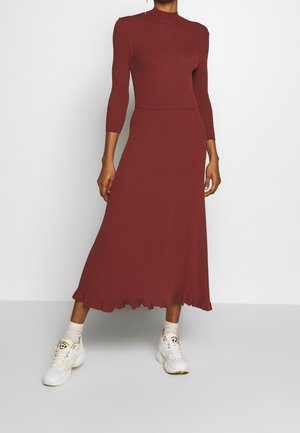 Personne portant une robe en maille côtelée de couleur rouille avec des manches trois-quarts et des baskets blanches avec des chaussettes beiges sur fond uni.