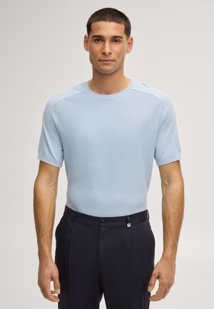 Mann trägt ein hellblaues, strukturiertes Kurzarmhemd, das in schwarze, hoch taillierte Hose gesteckt ist, stehend vor einem einfarbigen Hintergrund.