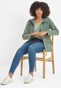 Light green waterproof jacket with a hood, paired with a light blue t-shirt and blue jeans. White sneakers with a subtle logo. Wooden chair.