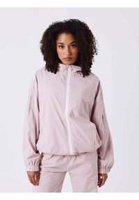 Chaqueta cortavientos rosa claro con cuello alto y cremallera completa en la parte delantera. Presenta puños elásticos y ribete blanco a lo largo de las costuras.